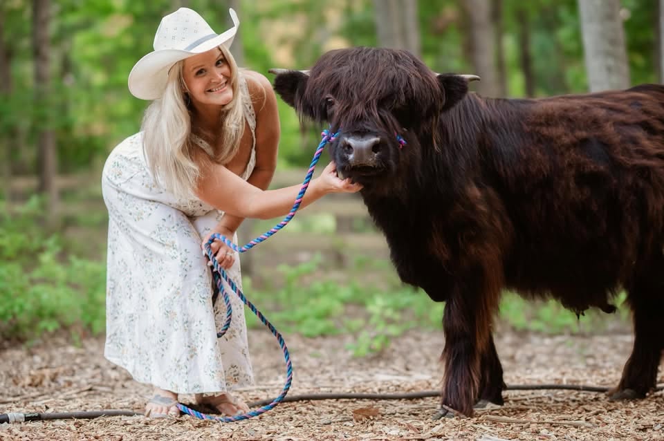 A smiling woman in a white hat beside a Highland cow